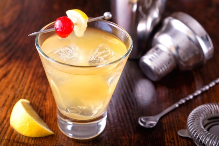 glass of whiskey sour cocktail garnished with cherry and a cocktail shaker